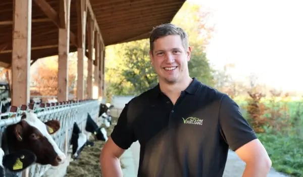 Porträt des lächelnden Landwirts Stefan Vogelsang in einem schwarzen Poloshirt mit dem Logo 'Hof Vogelsang'; im Hintergrund ist ein moderner Außenklimastall mit fressenden Milchkühen am Fressgitter zu sehen.