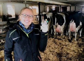 Eine lächelnde Tierärztin mit Brille steht im Kuhstall vor mehreren schwarzbunten Milchkühen und hält Blutprobenröhrchen in ihrer behandschuhten Hand.