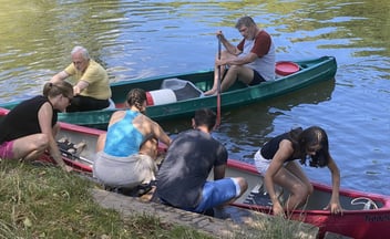 Eine Gruppe von Menschen bereitet sich am Flussufer auf eine Kanutour vor; zwei grüne und ein rotes Kanu liegen im Wasser, während die Teilnehmer einsteigen und die Boote startklar machen.