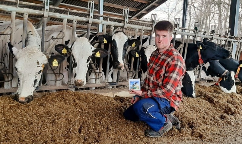 Landwirt Hillmann kniet im Kuhstall neben dem Futtertisch vor einer Reihe schwarzbunter Milchkühe und hält eine Produktverpackung des PerformaNat EnergieBolus in der Hand.