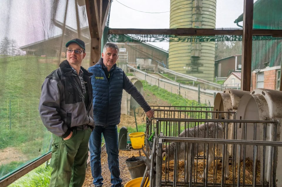 Ein Mitarbeiter von PerformaNat im fachlichen Austausch mit Michael Rentschler. Sie stehen unter einem Vordach neben Kälberiglus mit Stroheinstreu, in denen ein junges Kalb zu sehen ist. Im Hintergrund erstrecken sich das grüne Hofgelände, Wirtschaftsgebäude und ein Futtersilo.