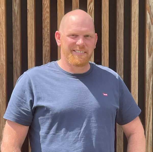 Christian Draht aus dem PerformaNat-Team blickt freundlich und lächelnd direkt in die Kamera. Der Mann mit Glatze und einem markanten, rötlichen Vollbart steht vor einer Wand aus vertikalen Holzlamellen und trägt ein schlichtes, graublaues T-Shirt mit einem kleinen Logo auf der Brust.