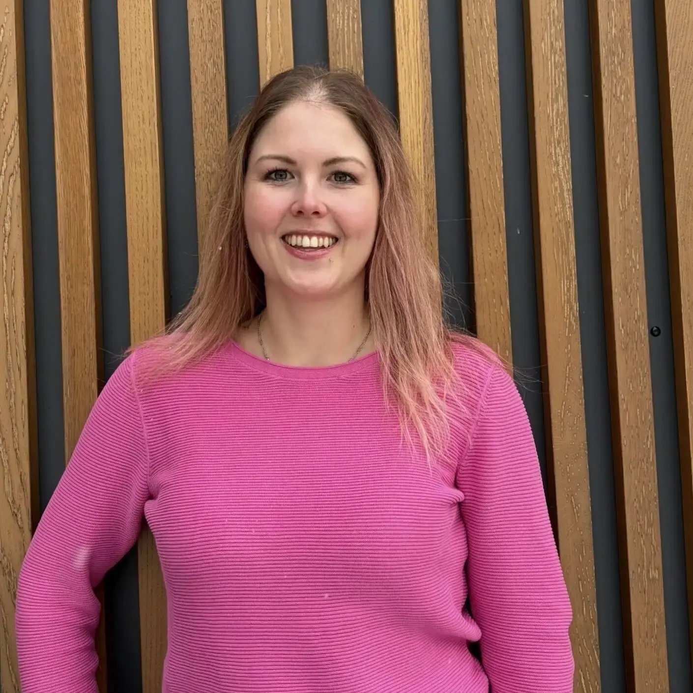 Diana Metten aus dem Bereich Kundenbetreuung bei PerformaNat strahlt Lebensfreude und Offenheit aus. Vor einer Wandgestaltung mit senkrechten Holzleisten zeigt sie sich mit mittellangen, rötlich-blonden Haaren und einem farbenfrohen, leuchtend rosafarbenen Feinstrickpullover.