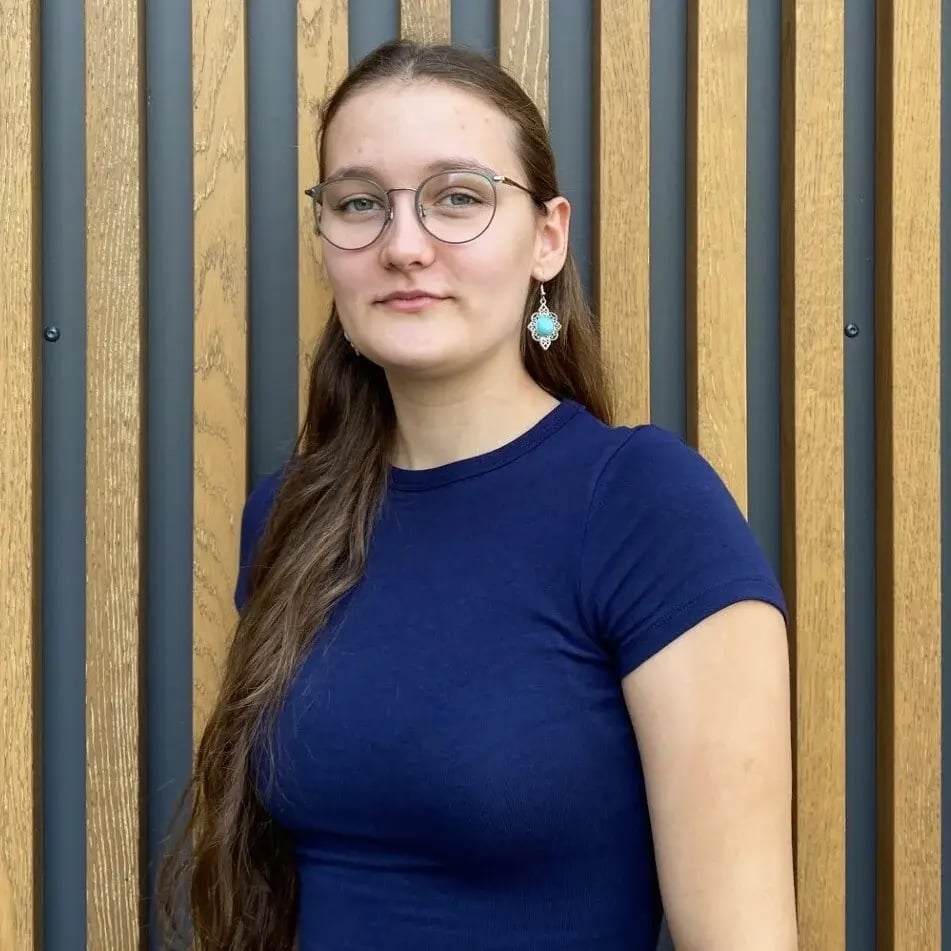 Hanna Tagmann aus dem PerformaNat-Team blickt mit einem ruhigen, freundlichen Gesichtsausdruck nach vorn. Sie hat langes braunes Haar, trägt eine runde Brille, auffällige Ohrringe mit hellblauen Schmucksteinen und ein schlichtes, dunkelblaues T-Shirt vor einer Fassade aus Holzlamellen.