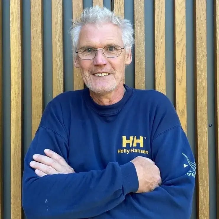 Helmut Staudinger aus dem PerformaNat-Logistikteam blickt mit einem offenen, freundlichen Ausdruck nach vorn. Er steht mit verschränkten Armen vor einer vertikalen Holzlamellenwand, trägt kurzes graues Haar, eine feine Brille und einen dunkelblauen Helly-Hansen-Pullover.