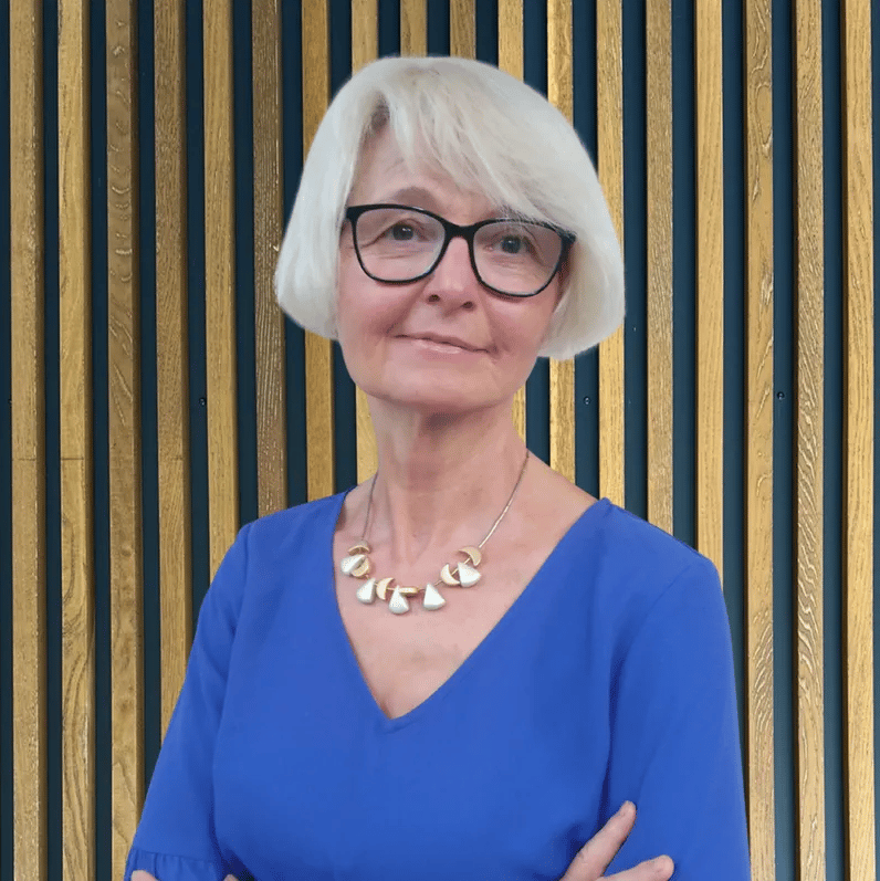 Ingrid Merimi aus der PerformaNat-Kundenbetreuung präsentiert sich mit sympathischem Blick vor einer modernen Holzfassade. Sie trägt eine markante schwarze Brille, eine helle Kurzhaarfrisur, ein königsblaues Oberteil sowie eine elegante Halskette.