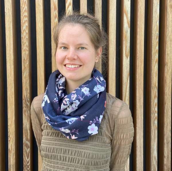 Janine Falke aus dem PerformaNat-Team blickt als Praktikantin freundlich und aufgeschlossen in die Kamera. Vor einem Hintergrund aus vertikalen Holzlamellen trägt sie ein hellbraunes, gerafftes Oberteil und einen dunkelblauen Loopschal mit einem dekorativen, floralen Muster aus hellen Blüten.