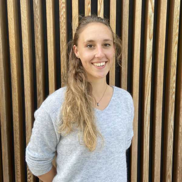 Leonie Urbaschik aus dem PerformaNat-Team lächelt herzlich und aufgeschlossen in die Kamera. Vor einer Wand aus vertikalen Holzlamellen trägt sie einen kuscheligen, hellgrauen V-Ausschnitt-Pullover und eine dezente Halskette.