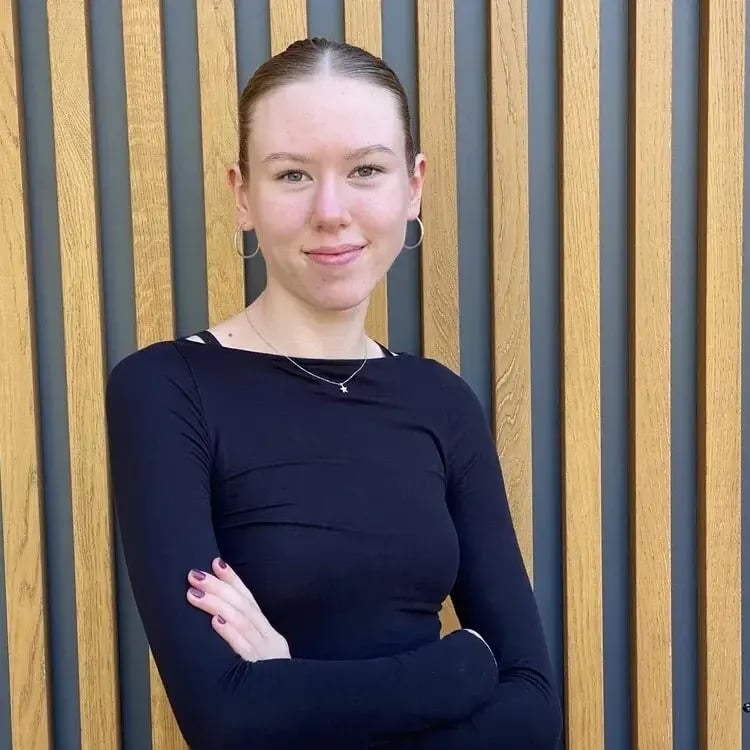 Ein Porträt von Lisann Jonscher, Werkstudentin bei PerformaNat. Sie hat braune, streng zurückgebundene Haare, trägt Creolen sowie eine Halskette und blickt lächelnd in die Kamera. Sie steht mit verschränkten Armen in einem schwarzen Oberteil vor einer Holzlamellenwand.