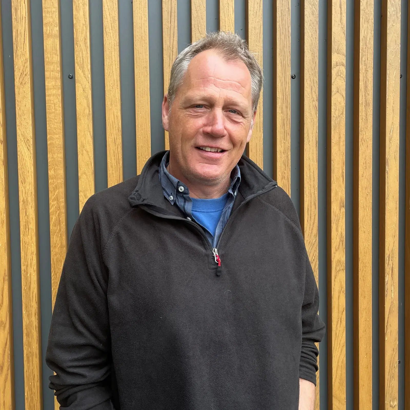 Roland Berger aus dem PerformaNat-Team blickt mit einem freundlichen Lächeln in die Kamera. Der Mann mit kurzen, leicht graumelierten Haaren steht vor einer Wand aus vertikalen Holzlamellen und trägt einen schwarzen Fleece-Pullover mit Reißverschluss über einem blauen Hemd und T-Shirt.