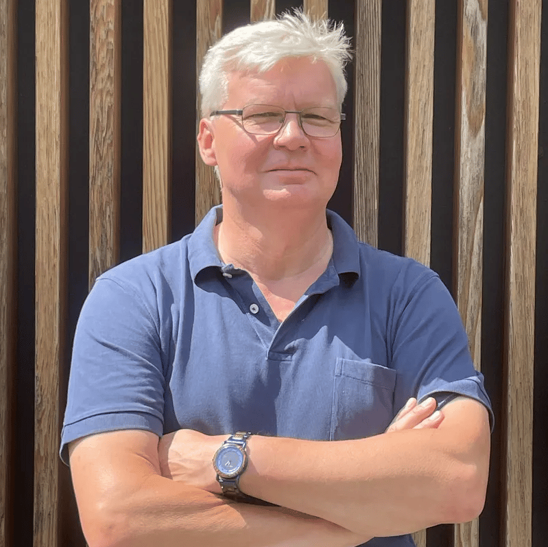 Stefan Andler aus dem PerformaNat-Team posiert mit verschränkten Armen und einem freundlichen, gelassenen Gesichtsausdruck vor einer stilvollen Wand aus vertikalen Holzlatten. Er trägt kurzes weißes Haar, eine dezente Brille, eine Armbanduhr und ein blaues Poloshirt.