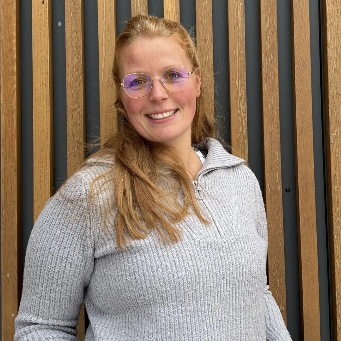 Wiebke Nünning aus der PerformaNat-Kundenbetreuung strahlt Offenheit und Herzlichkeit aus. Vor dem Hintergrund vertikaler Holzlamellen zeigt sie sich mit langen, rötlich-blonden Haaren, einer eleganten, filigranen Brille und einem gemütlichen, grauen Strickpullover mit Reißverschlusskragen.