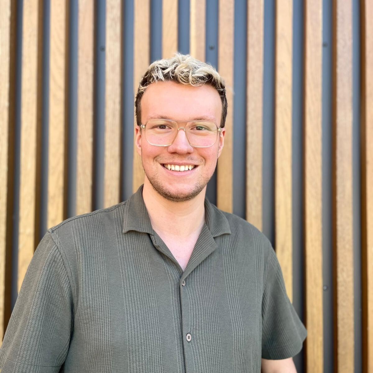 Portrait of Simon Schnieder, team member at PerformaNat, standing in front of a modern wooden paneled wall.