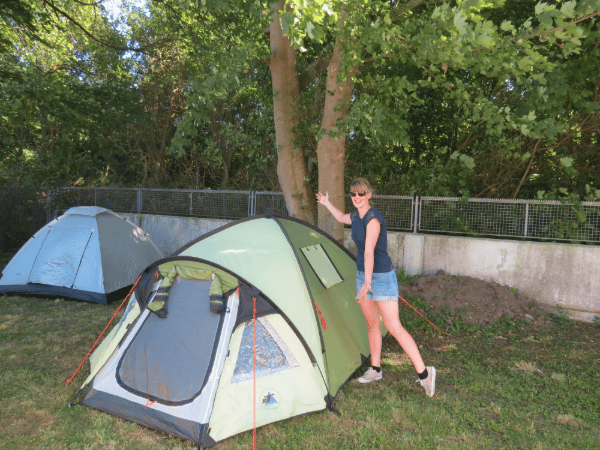Katharina Schrapers mit Sonnenbrille, dunkelblauem T-Shirt und kurzen Jeans-Shorts steht lächelnd auf einer grünen Wiese neben einem aufgebauten hellgrünen Campingzelt.