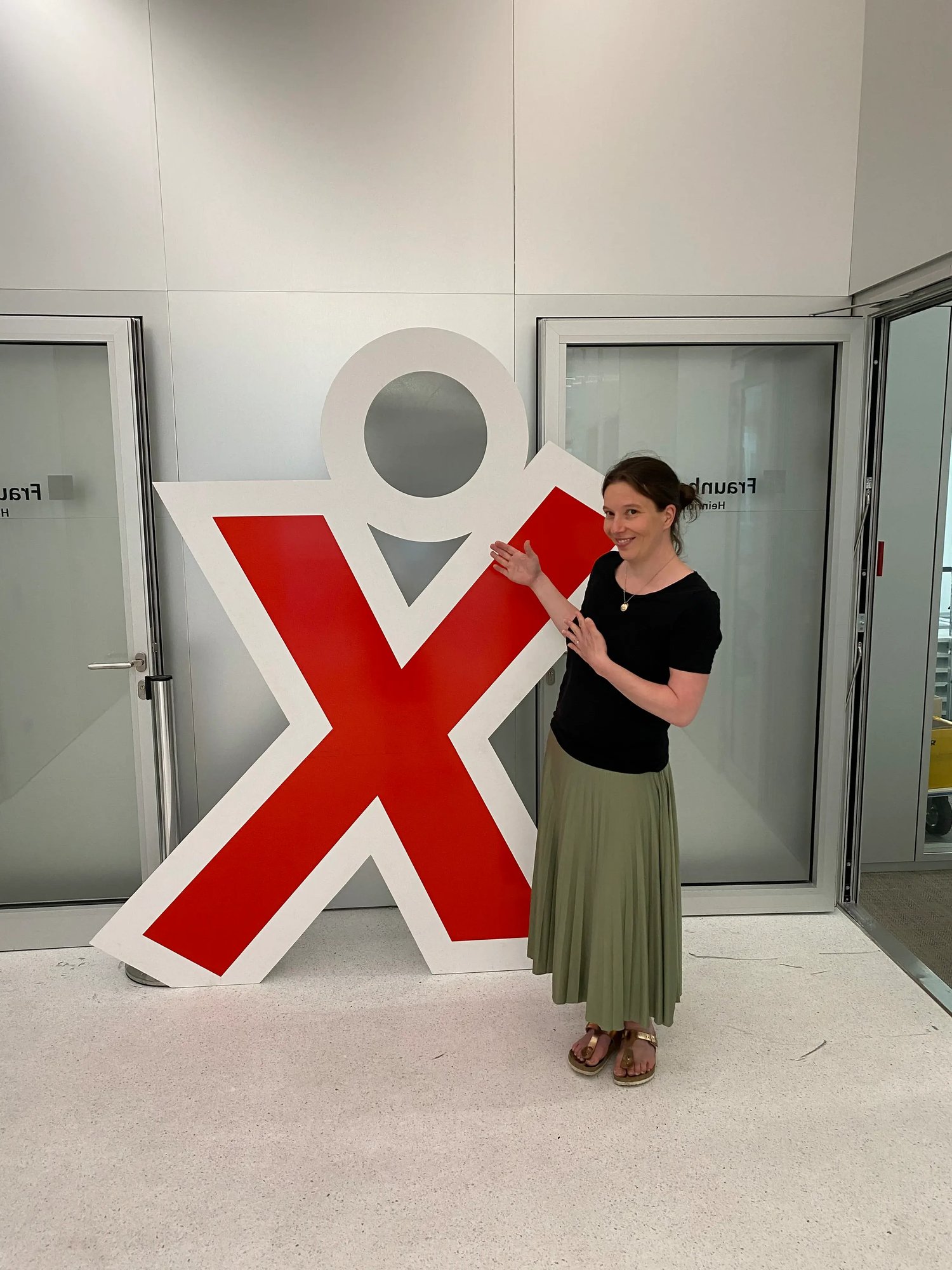 Julia Rosendahl steht in einem modern gestalteten Innenraum neben einer großen Aufstellfigur in Form eines roten 'X' mit einem Kreis als Kopf. Sie trägt ein schlichtes schwarzes T-Shirt, einen langen, olivgrünen Rock und Sandalen.