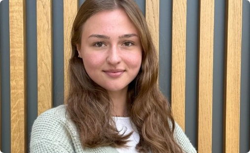 Porträt einer lächelnden jungen Frau mit langen braunen Haaren, die einen hellgrünen Strickcardigan und ein weißes T-Shirt trägt, vor einer vertikalen Holzlattenwand.
